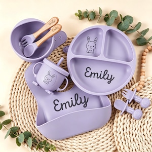 May include: A lavender baby feeding set featuring a divided plate, bowl, bib, cup, and utensils. The set includes a spoon and fork with wooden handles, and the name "Emily" is printed on the plate and bib. A cute bunny illustration is on the plate and cup.