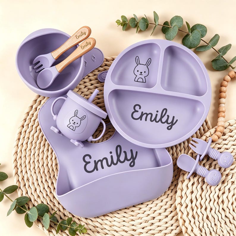 May include: A lavender baby feeding set featuring a divided plate, bowl, bib, cup, and utensils. The set includes a spoon and fork with wooden handles, and the name "Emily" is printed on the plate and bib. A cute bunny illustration is on the plate and cup.