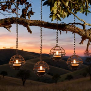 May include: Four hanging candle holders with lit candles suspended from a tree branch. The spherical holders are made of a dark metal cage design, and the background features a sunset over rolling hills.