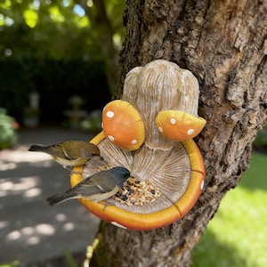 Könnte beinhalten: Ein pilzförmiger Vogelfutterautomat, der an einem Baumstamm befestigt ist. Der Futterautomat ist in Orange und Weiß bemalt. Zwei kleine Vögel fressen Samen. Ein bezaubernder Gartenschmuck, um Wildtiere anzulocken.