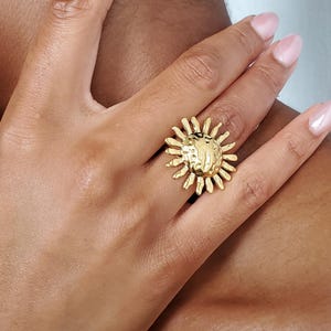 May include: A gold-toned ring featuring a central circular disc with a hammered, textured surface, surrounded by radiating thin, pointed rays that mimic the appearance of a sun. The metal has a polished finish that reflects light.