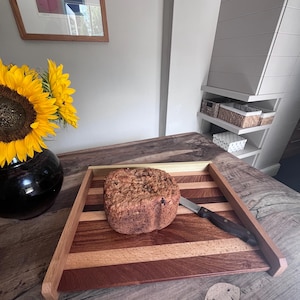 May include: A square wooden serving tray featuring alternating light and dark wood stripes, a raised border, and a metallic accent edge. A loaf of brown bread and a stainless steel knife with a black handle are placed on the tray surface.