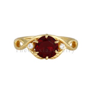 May include: A gold ring featuring a large, round, deep red garnet gemstone at the centre, flanked by two small, clear, round-cut accent stones. The band has an infinity-style twisted design. The text TheGixiaJewelry is visible across the centre.