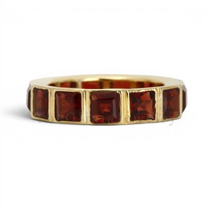 Puede incluir: Un anillo de banda de oro que presenta una serie de piedras preciosas de granate rojo oscuro, de corte cuadrado, colocadas una al lado de la otra a lo largo de toda la circunferencia. El metal tiene un acabado pulido y las piedras están sujetas por barras.