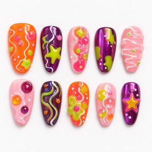 May include: A collection of ten colorful, decorated artificial nails. Designs include orange, purple, and pink bases with various patterns like flowers, stars, and swirls. The nails are adorned with raised, glossy details in contrasting colors.