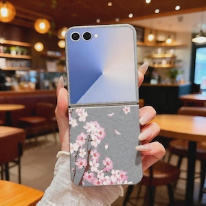 Può includere: Una custodia per telefono grigia e blu con un design floreale. La custodia è aperta, rivelando le lenti della fotocamera del telefono. La metà inferiore della custodia presenta uno sfondo grigio con rami di fiori di ciliegio rosa e petali che cadono.