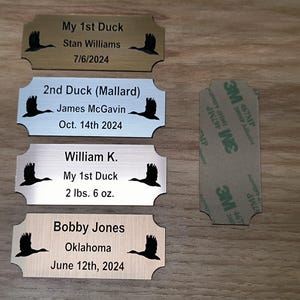 May include: Four engraved metal nameplates with a duck silhouette, each with personalized text. One plate reads "My 1st Duck" and includes a weight of 2 lbs 6 oz. A 3M adhesive pad is also visible.