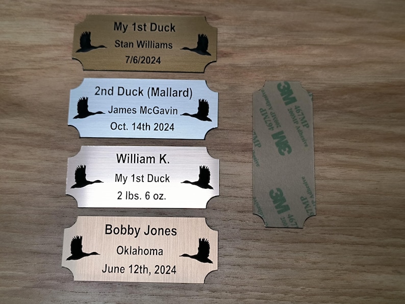 May include: Four engraved metal nameplates with a duck silhouette, each with personalized text. One plate reads "My 1st Duck" and includes a weight of 2 lbs 6 oz. A 3M adhesive pad is also visible.