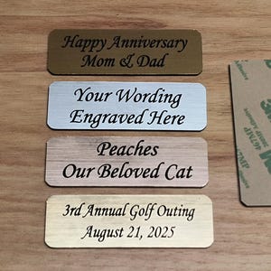 May include: A collection of rectangular engraved metal nameplates in various colors, including gold, silver, and wood-tone. The plates feature personalized text such as "Happy Anniversary Mom & Dad" and "Peaches Our Beloved Cat."