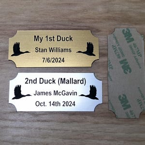 May include: Two metal plaques with a duck silhouette and text. One is gold-toned, reading "My 1st Duck" with a name and date. The other is silver, labeled "2nd Duck (Mallard)" with a name and date. A 3M adhesive strip is also present.