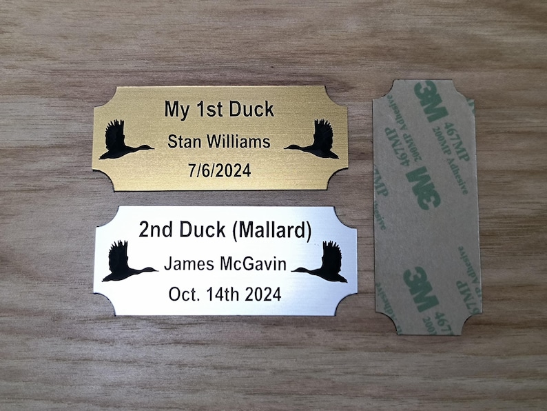 May include: Two metal plaques with a duck silhouette and text. One is gold-toned, reading "My 1st Duck" with a name and date. The other is silver, labeled "2nd Duck (Mallard)" with a name and date. A 3M adhesive strip is also present.