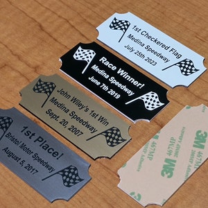 May include: Four silver, gold, black, and white award plaques with checkered flags and text that reads "1st Checkered Flag Medina Speedway July 25th 2023", "Race Winner! Medina Speedway June 7th 2019", "John Wiley's 1st Win Medina Speedway Sept. 20, 2007", and "1st Place! Bristol Motor Speedway August 5, 2017".