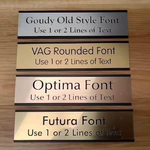 May include: Four gold and silver metal plaques with black trim. Each plaque has a different font style and the text "Use 1 or 2 Lines of Text". The fonts are Goudy Old Style, VAG Rounded, Optima, and Futura.