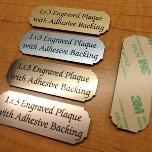 May include: Four 1x3 inch engraved plaques with adhesive backing. The plaques are made of gold and silver metal. The text on the plaques reads "1x3 Engraved Plaque with Adhesive Backing".