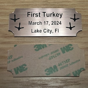 May include: A rectangular, copper-colored metal plaque with rounded corners. The plaque reads "First Turkey" above "March 17, 2024" and "Lake City, FI" in black text. Black turkey footprints are on each side. A 3M adhesive backing is below.