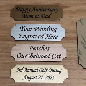 May include: Four engraved metal plaques with black text. The plaques include phrases like "Happy Anniversary Mom & Dad", "Your Wording Engraved Here", "Peaches Our Beloved Cat", and "3rd Annual Golf Outing August 21, 2025".
