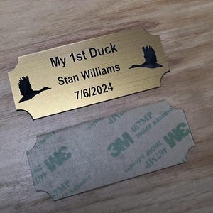 May include: A gold-colored metal plaque with the text "My 1st Duck Stan Williams 7/6/2024" and two black duck silhouettes. A 3M adhesive backing is visible below the plaque. The plaque is on a wooden surface.