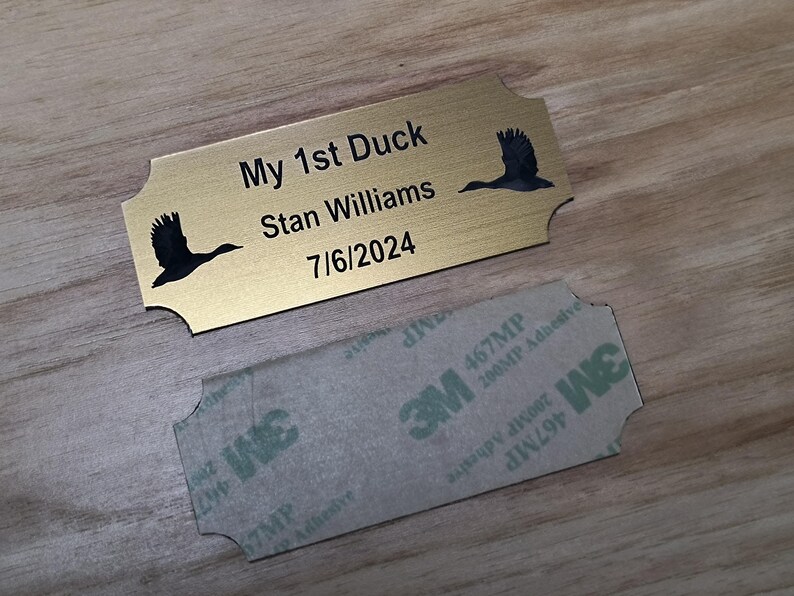 May include: A gold-colored metal plaque with the text "My 1st Duck Stan Williams 7/6/2024" and two black duck silhouettes. A 3M adhesive backing is visible below the plaque. The plaque is on a wooden surface.