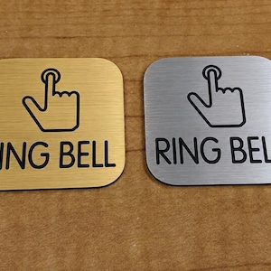 May include: Two square metal doorbell signs, one gold and one silver, with rounded corners. Each sign features a black outline of a finger pressing a button above the words "RING BELL" in black, sans-serif font.