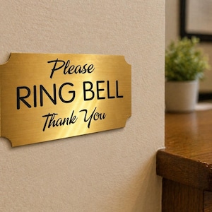May include: A rectangular brass-colored sign with the words "Please RING BELL Thank You" in black script. The sign is mounted on a light-colored wall, with a wooden desk and plant in the background.