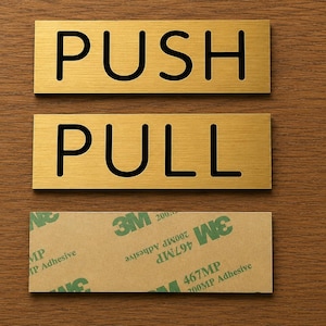 May include: Two rectangular gold door signs with black text. The top sign says "PUSH", the bottom sign says "PULL". Below the signs is a rectangular adhesive strip.