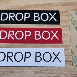 May include: Three rectangular signs with the text "DROP BOX" in white lettering on black, red, and white backgrounds. A strip of 3M adhesive is next to the signs. The signs are on a wooden surface.