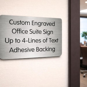 May include: A rectangular, brushed metal office suite sign with rounded corners. The sign reads "Custom Engraved Office Suite Sign Up to 4-Lines of Text Adhesive Backing" in black text. The sign is mounted on a light-colored wall.