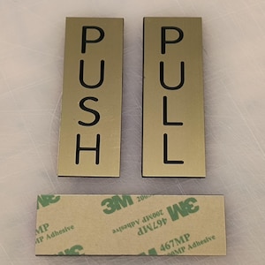 Peut inclure: Deux panneaux métalliques dorés avec des lettres noires. Le panneau de gauche indique "PUSH" et le panneau de droite indique "PULL".