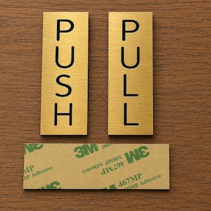 May include: Two rectangular gold-colored door signs with black lettering. One sign reads "PUSH" and the other reads "PULL". A rectangular adhesive strip is below the signs. The signs are on a brown wooden surface.