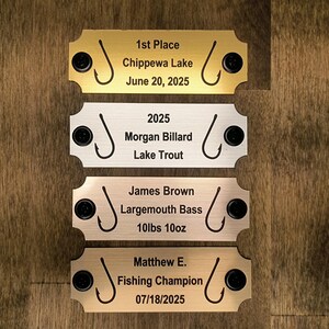 May include: Four metal fishing awards with black accents and text. The top award is gold and reads "1st Place, Chippewa Lake, June 20, 2025." The second is silver and reads "2025, Morgan Billard, Lake Trout." The third is bronze and reads "James Brown, Largemouth Bass, 10lbs 10oz." The bottom award is gold and reads "Matthew E., Fishing Champion, 07/18/2025."