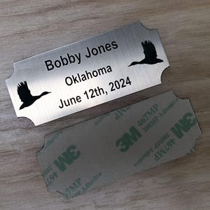 May include: A silver metal nameplate with rounded edges, featuring the name "Bobby Jones", the location "Oklahoma", and the date "June 12th, 2024". Black silhouettes of flying birds are on the plate. A 3M adhesive backing is visible.