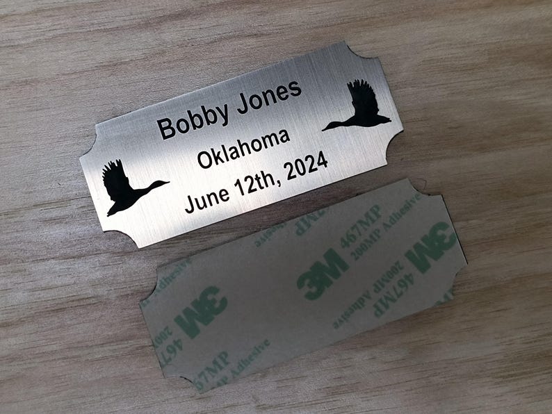 May include: A silver metal nameplate with rounded edges, featuring the name "Bobby Jones", the location "Oklahoma", and the date "June 12th, 2024". Black silhouettes of flying birds are on the plate. A 3M adhesive backing is visible.