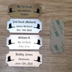 May include: Four personalized metal nameplates with a duck silhouette. Text includes names, dates, and weights. One plate is gold-toned, two are silver-toned, and one is wood-toned. A 3M adhesive strip is on the right.