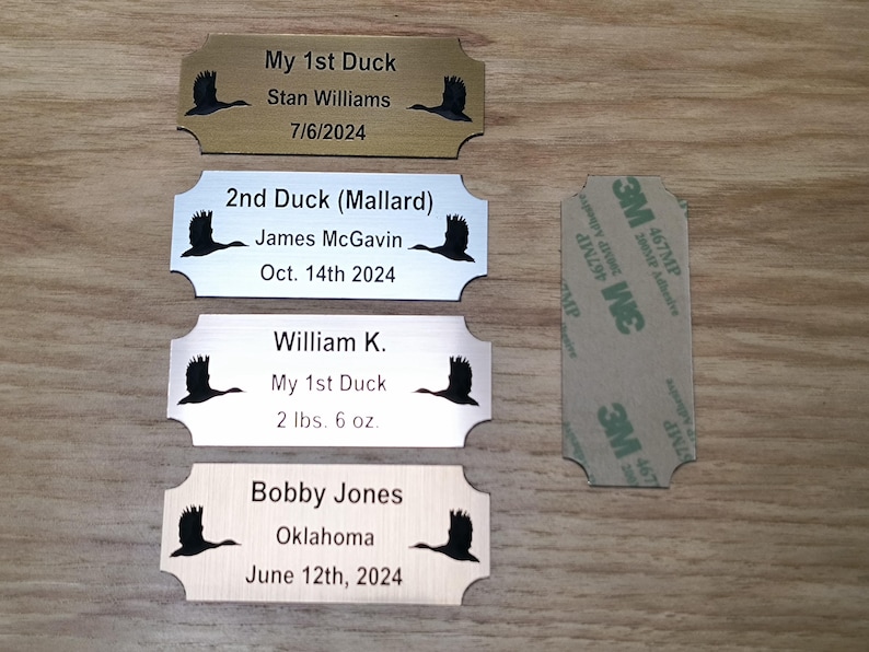 May include: Four personalized metal nameplates with a duck silhouette. Text includes names, dates, and weights. One plate is gold-toned, two are silver-toned, and one is wood-toned. A 3M adhesive strip is on the right.