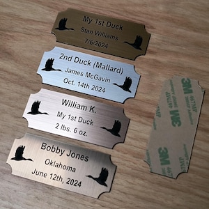 May include: Four metal nameplates with a duck silhouette and engraved text. The plates include names, dates, and details like "My 1st Duck" and weights in pounds and ounces. A 3M adhesive pad is also visible.