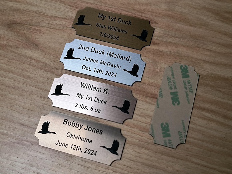 May include: Four metal nameplates with a duck silhouette and engraved text. The plates include names, dates, and details like "My 1st Duck" and weights in pounds and ounces. A 3M adhesive pad is also visible.