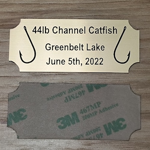 May include: Gold-colored metal plaque with a black inscription that reads "44lb Channel Catfish Greenbelt Lake June 5th, 2022". The plaque has two black fishing hooks on either side of the text.