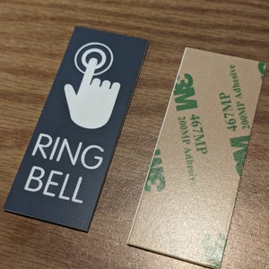 May include: Rectangular sign with the words "RING BELL" in white text, and a white hand pressing a button icon on a dark gray background. A separate adhesive strip is next to the sign.