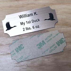 May include: A brushed metal nameplate with the text "William K.", "My 1st Duck", and "2 lbs. 6 oz." Two black duck silhouettes are on the plate. A 3M adhesive backing is below the plate.
