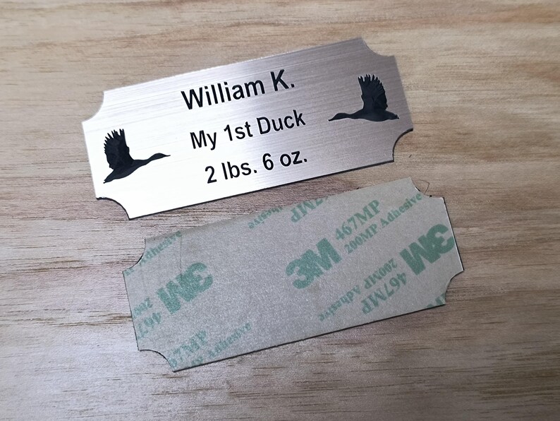May include: A brushed metal nameplate with the text "William K.", "My 1st Duck", and "2 lbs. 6 oz." Two black duck silhouettes are on the plate. A 3M adhesive backing is below the plate.