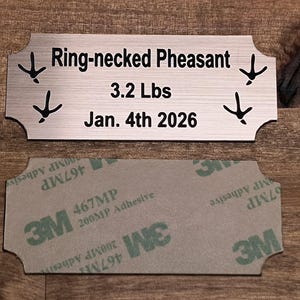 May include: A brushed metal sign with the text "Ring-necked Pheasant", "3.2 Lbs", and "Jan. 4th 2026". The sign has decorative bird footprints and is shown with its adhesive backing.