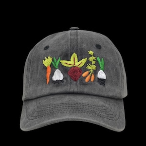 Peut inclure: Casquette de baseball grise avec broderie de légumes. La conception comprend une carotte, de l'ail, une betterave et d'autres légumes en orange, vert, jaune et rouge. La casquette a une visière incurvée.