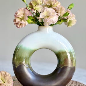 May include: A circular ceramic vase with a smooth, glossy finish featuring a gradient color scheme that transitions from white at the top to olive green in the middle and dark brown at the base, filled with a bouquet of light pink carnations.