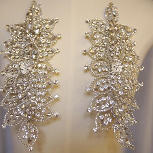 May include: A pair of elongated, symmetrical appliques featuring a dense arrangement of clear, faceted crystals set in silver-toned metal. The design consists of multiple leaf-shaped segments layered to create a textured, shimmering floral pattern.
