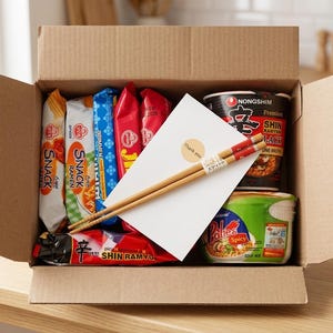 May include: A cardboard box filled with various instant ramen products, including cups and packets. A white card with a sticker and a pair of chopsticks rest on top. The box is open, revealing the contents.