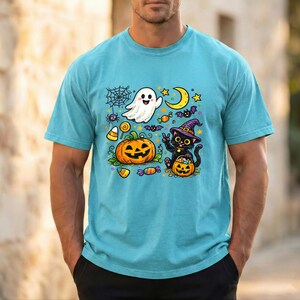 May include: A light blue t-shirt featuring a colorful Halloween-themed graphic print. The design includes a white ghost, a black cat wearing a purple witch hat, a jack-o'-lantern, a crescent moon, a spider web, and various pieces of candy.