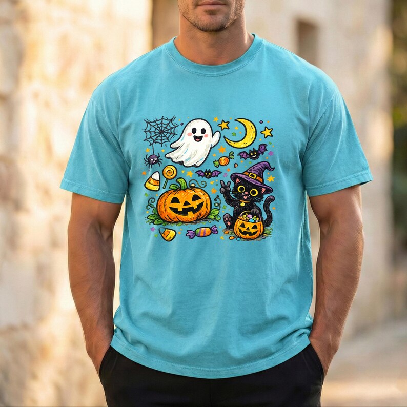 May include: A light blue t-shirt featuring a colorful Halloween-themed graphic print. The design includes a white ghost, a black cat wearing a purple witch hat, a jack-o'-lantern, a crescent moon, a spider web, and various pieces of candy.