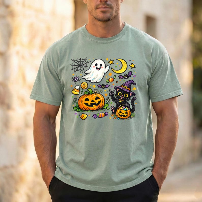 May include: A sage green short-sleeve t-shirt featuring a colorful Halloween graphic print. The design includes a white ghost, a jack-o'-lantern, a black cat wearing a purple witch hat, a yellow crescent moon, spiderwebs, and assorted candies.