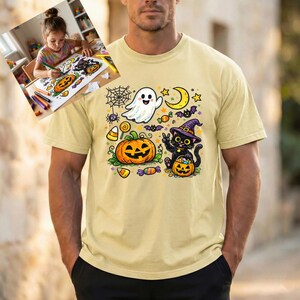 May include: A light yellow short-sleeve t-shirt featuring a colorful Halloween graphic print. The design includes a white ghost, a large orange jack-o'-lantern, a black cat wearing a purple witch hat, a yellow crescent moon, and candy.