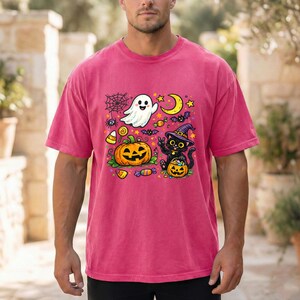 May include: A pink short-sleeve t-shirt featuring a colorful Halloween graphic. The design includes a white ghost, a large orange jack-o'-lantern, a black cat wearing a purple witch hat, a yellow crescent moon, bats, candy, and a spider web.